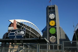 The interior of a stadium from the upper tier behind the south end zone during the day. The end zones and seating sections are colored blue. At the north end is a smaller seating area at the base of a tower. Several high-rise office buildings are in the distance.