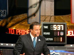 Candid photograph of Mariucci seated behind a desk on a TV set wearing a dark pin-striped suit, blue striped tie and a headset