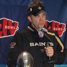 Color head-and-shoulders photograph of white man (Sean Payton) wearing a black New Orleans Saints jacket and a black Saints visor, and holding a microphone in front of a blue Super Bowl XLIV backdrop.