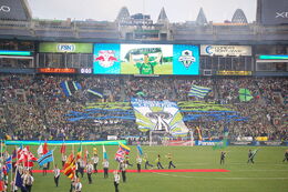 A stand of a stadium with fans holding a large banner depicting the Space Needle. Green and blue flags wave throughout the stand and the flags of different nations are held by people on the field.