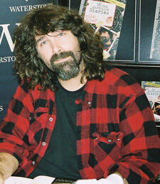 A male with medium-length hair and a goatee, wearing a red-and-black plaid buttoned shirt on top of a black t-shirt poses at an autograph signing.