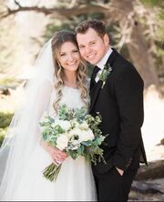 Jeremiah and Hannah standing together and smiling at the camera. Hannah is wearing a white dress and veil and is holding a green and white bouquet