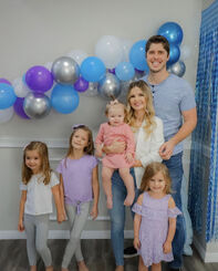 John, Alyssa and their four daughters outside on the 4th July. John and Alyssa are both crouched down, John is wearing a blue suit with white shirt and red tie, and Alyssa is wearing a red dress and nude heels. John's right arm (at the left of the photo) is around Lexi, who is leaning against his side. Allie is sitting on his other knee. Zoey is standing on the other side of Alyssa, and Maci is held on Alyssa's lap. the three older girls are wearing a white dress with a unicorn on the chest, and red white and blue tulle skirt. they also have a red bow in their hair and Allie is holding a pink clutch. Maci is wearing a frilly white patterned romper and has a blue bow on her head.