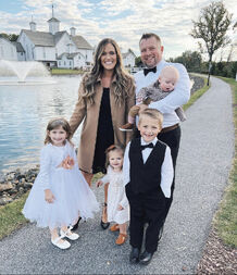 Whitney is wearing a blue shirred dress, and is holding her bump. Next to her stands Zach (in a dark suit), holding Khloé (in a white dress with her hair in a blue bow). In front of them stands Bradley (in a black jacket, blue shirt and black bow tie, and beige trousers) linking arms with Kaci (in a white dress with a blue bow in her hair.