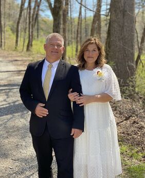 Gil and Kelly standing in a wood. Gil is wearing a dark suit with a white shirt and Kelly is wearing a white dress with a flower on