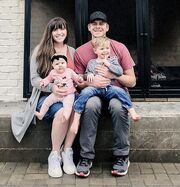 Joy and Austin sitting on a ledge. Joy is wearing a blue denim skirt, orange shirt and grey cardigan. She has Evelyn on her lap who has a dark bow on her head and a pink top and leggings. Next to Joy is Austin, who has a black baseball cap on, is wearing a dark pink/faded red shirt and black trousers. He has Gideon on his lap, who is smiling and is wearing a grey shirt and denim jeans.