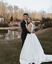 Travis and Katie standing with their arms around each other on their wedding day. Travis is wearing black, and Katie is wearing a long, empire, wedding dress.