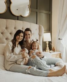Carlin and Evan sitting on their bed, with Evan holding Layla on his lap and Carlin holding Zade in her arms. Layla has two little bunches on the top of her head, all of the family are wearing neutral coloured clothing, and all but Zade (who is asleep and has chubby cheeks) are smiling at the camera.