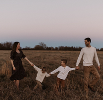 the Dillard family, walking through a field, Jill on the left wearing a dark dress. Jill is holding hands with Sam, who is holding hands with Israel, and Israel is holding hands with Derick. Derick and the two boys are wearing pale/white tops and brown trousers.