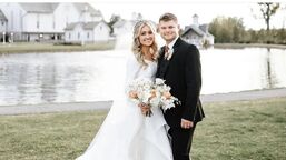 Esther, wearing a white dress and a tiara, holding a bouquet of white and pink flowers, and Nathan, wearing a black suit, are standing in front of a large pond smiling at the camera.