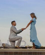 Nathan, down on one knee and wearing a grey suit, is holding the hand of Esther, who is smiling down at him. She is wearing a long blue flowey dress.