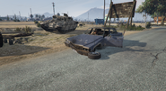 A Rhino Tank in GTA V, with an Emperor that has been totalled by having the Rhino drive over it.