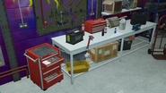 A worktable featuring a car battery, wrench, several oil containers, a blow torch, a Power Metal tool box, an exhaust with silencer/catalyst, a Power Metal drill case, Power Metal tool chest, and a vice.