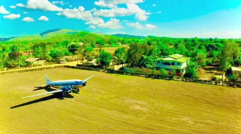A vintage twin-engine aircraft on the grass runway at Pignon Airport, with the town’s homes and northern hills in the background.