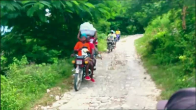 Passengers and goods traveling by motorcycle along an unpaved hillside road in Goyavier.