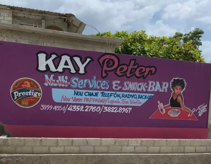 Kay Peter Multi-Services & Snack Bar, a neighborhood spot near Barbe offering phone charging, radio and backup services, mobile payments, and quick snacks—typical of the small roadside businesses that serve daily needs in the Hauts-de-Saint-Marc.