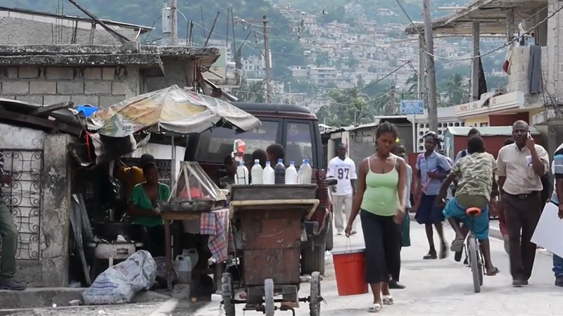 Life moves steadily through the narrow streets of Cité de Dieu — vendors at their stalls, children weaving past on bicycles, and neighbors carrying water beneath the hills that rise behind Port-au-Prince.