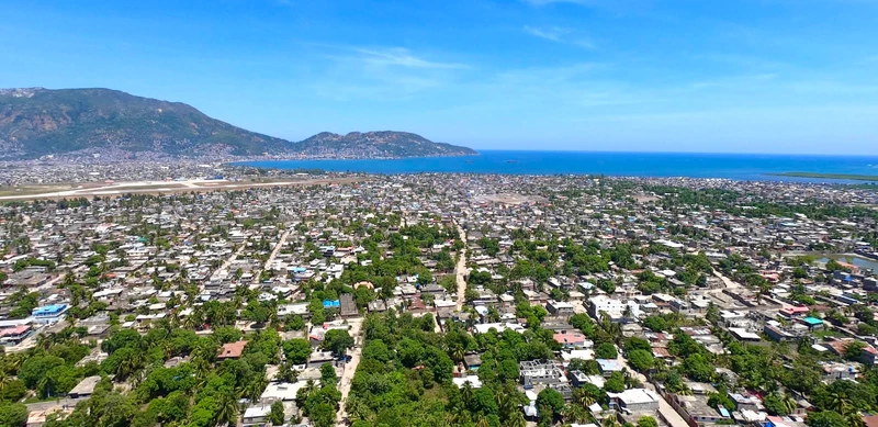 An aerial view over , looking north toward the bay and the mountains of  and Morne du Cap, with the runway of Hugo Chávez International Airport visible on the left and the dense grid of Petite-Anse stretching toward the coast.