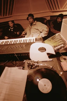 Hip hop producer and rapper RZA in a music studio with two collaborators. Pictured in the foreground is a synthesizer keyboard and a number of vinyl records; both of these items are key tools that producers and DJs use to create hip hop beats.
