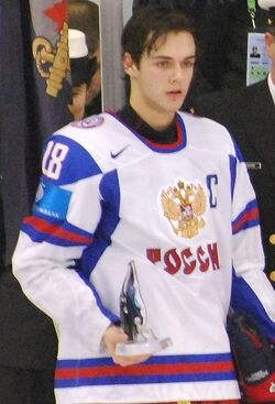 A young man with dark hair wearing a white jersey holds a small trophy in his right hand.