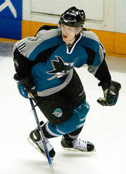 An ice hockey player skating in mid-stride. He holds his stick with one hand along the ice while looking to his right. He wears a teal jersey with black trim, as well as a black, visored helmet.