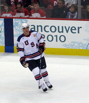 An ice hockey player is skating on the ice. He is in a white jersey with the letters "USA" going diagonally across his jersey.