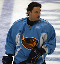 An ice hockey player is standing while slightly turned to his left. He has short dark hair and is not wearing a helmet. He is wearing a blue uniform with a large orange bird with a ice hockey stick on his chest.