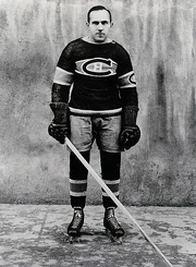Full profile of a balding ice hockey player in full uniform leaning forward on his stick with a serious look on his face.