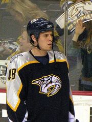 An ice hockey player looking to the right of the camera. He is wearing a black helmet and a black uniform with a large sabertooth head on his chest.