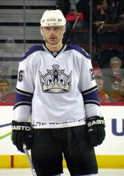 An ice hockey player standing facing towards the camera. He is wearing a white helmet, and a white, black, and purple uniform with a large crown on his chest.