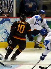 An ice hockey player dressed in a black and orange jersey watchfully skates in front of an opposing player who is playing the puck.