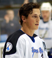 Upper body of a man with short black hair as he stares into the distance. He is in a white uniform with black trim.