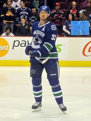 An ice hockey player wearing a blue and green jersey and a blue, visored helmet. He is following through with his hockey stick while standing upright.