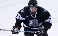 Young male ice hockey player in full gear, including a face mask, skates toward the camera with a determined expression as he looks past the camera.