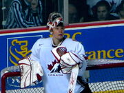 Hockey player in white uniform and goaltender's uniform. He leans against a goal, his helmet lifted above his head, and looks upward.