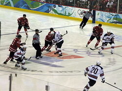 An ice hockey game. Two referees in black and white striped shirts are visible, as are four players from each team. One team is wearing red jerseys with a maple leaf on the front, and the other is wearing white jerseys with blue accents and the letters "U-S-A" on the front.