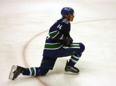 An ice hockey player dressed in a blue jersey with blue and green trim. He is stretching with one knee on the ice and the other lunged forward. He rests his stick on one leg while looking forward.