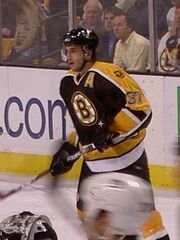 Hockey player in black and yellow uniform with a large "B" in the middle. His mouth is slightly open and his stick is raised diagonally off the ground.