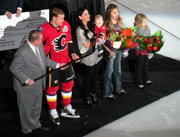 A man in a red hockey uniform accepts a silver stick from another man in in a grey suit as his wife and three young daughters stand beside.