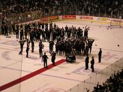The members of an ice hockey team collectively raise a trophy in celebration while numerous dignitaries and members of the media surround them.