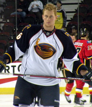 An ice hockey player stands directly upright holding an ice hockey stick horizontally across his stomach. He is wearing a no helmet and is wearing a black and white uniform with a large orange bird with a ice hockey stick on his chest.