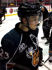 Young male ice hockey player, visible from waist up, skating while facing the right. He is wearing a helmet with a visor, and the uniform logo, a snarling moose, is partially visible on his jersey. In the background three of his teammates, partially obscured, are also warming up.