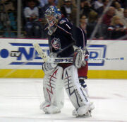 Hockey player in dark blue uniform and goaltender's gear. He stands on the ice with his hands on thighs; the left hand holds a hockey stick parallel to the rink.