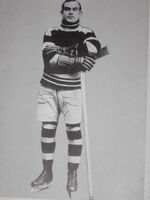 a black and white photo of a young hockey player standing on his skates with his arms folded leaning slightly on an upside down hockey stick