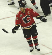 A hockey player in a red and black uniform with a stylized C logo on his chest skates across the ice.