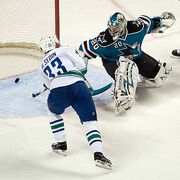 An ice hockey player wearing a white and blue jersey scores against a goaltender wearing a teal and black jersey with white pads. The player has both hands on his stick outstretched to direct the puck in the net, while the goaltender is off balance looking back at the puck in his net.