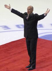 A bald man in a black suit raises his arms to acknowledge a crowd that is not seen.