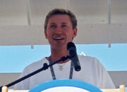 Upper body of a man with dirty-blonde hair who is looking into the distance. He is standing at a podium and speaking into a microphone.