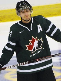 A Caucasian ice hockey player in his late teens wearing a green jersey with a red and black logo of a stylized maple leaf.