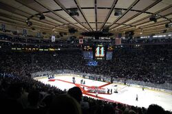 An arena set up for ice hockey. A red carpet sits atop the ice, with a group of people in white jerseys on it.